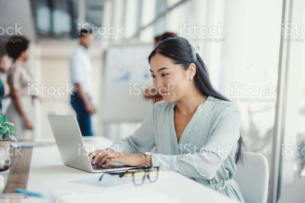 Asian woman working on laptop. Businesswoman busy working on laptop computer at office with colleagues in the background.